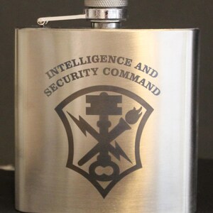 May include: A stainless steel flask with a silver cap. The flask features the words "INTELLIGENCE AND SECURITY COMMAND" in a curved font above a black emblem with a key, lightning bolt, and torch.