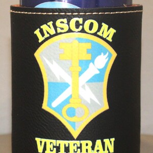 May include: Black neoprene can cooler with a yellow and blue insignia that says "INSCOM VETERAN".