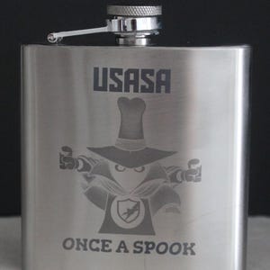 May include: A stainless steel flask with a hinged cap. The flask features the text "USASA" above an etched image of a cartoon figure in a hat and cape, with the words "ONCE A SPOOK" below.