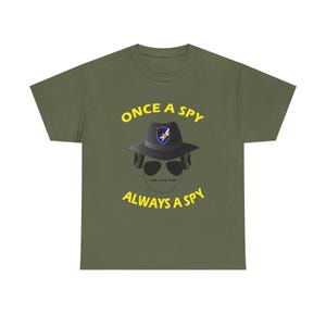May include: Olive green t-shirt with the text "ONCE A SPY ALWAYS A SPY" in yellow. The graphic features a spy wearing a fedora, sunglasses, and headphones.