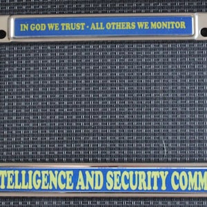 May include: A chrome license plate frame with blue and yellow text that reads "IN GOD WE TRUST - ALL OTHERS WE MONITOR" and "INTELLIGENCE AND SECURITY COMMAND". The frame has two mounting holes.