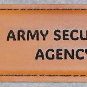 May include: A flat, rectangular metal bottle opener with a tan leather-like wrap around the center. The leather features a black printed shield emblem with a lightning bolt graphic and the text ARMY SECURITY AGENCY in bold black lettering.