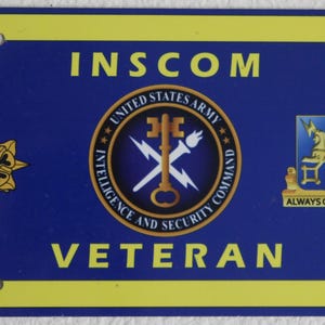 May include: Blue license plate with yellow trim featuring the text "INSCOM VETERAN." Includes the United States Army Intelligence and Security Command emblem, a gold floral emblem, and a crest with the words "ALWAYS OUT FRONT."