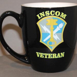May include: Black ceramic mug with a white interior. The mug features the text "INSCOM VETERAN" in yellow, along with a shield emblem with a key, lightning bolts, and a torch.
