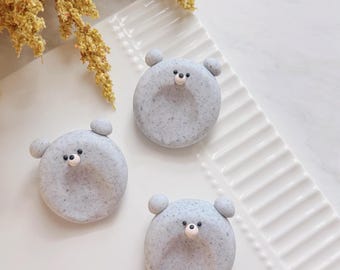 May include: Three circular, light gray speckled bear-shaped objects with small rounded ears and a central indentation. Each bear features a small, light-colored snout with a black dot for a nose. The items are arranged on a white surface.