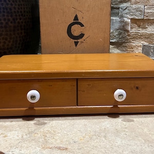 May include: A rectangular wooden storage box with a medium brown finish, featuring two side-by-side drawers. Each drawer is equipped with a small, round, white ceramic knob with a metallic center. The box has a smooth, flat top surface.