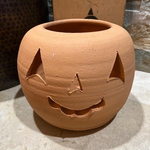 May include: A terracotta pumpkin-shaped planter with a carved face. The pumpkin has triangular eyes, a triangular nose, and a curved mouth. The planter is round with a wide opening at the top. The color is a warm orange-brown.