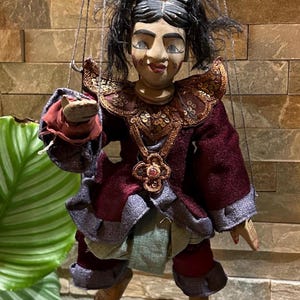 May include: A vintage marionette doll with a painted wooden face and black hair in pigtails. The doll wears a burgundy and gray outfit with gold sequin accents. The puppet is suspended by strings, showcasing its intricate design and craftsmanship.