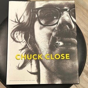 May include: A book titled "CHUCK CLOSE" featuring a black and white portrait of a man wearing glasses and smoking a cigarette. The book cover has a light background and the text is in yellow.