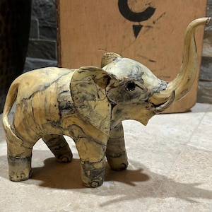 May include: A small, decorative elephant figurine with a raised trunk. The elephant is primarily cream-colored with black marbling throughout. The figurine has large ears and small tusks. The elephant is standing on four legs.