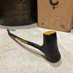 May include: A handcrafted, black wooden pipe with a natural bark texture. The bowl is painted black with a yellow top. The stem is thin and dark, with a small label that reads "Irish shillelagh". A green string is attached.