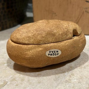 May include: A tan, oval-shaped ceramic potato baker with a textured, stone-like exterior. The lid sits atop the base, and a small white oval sticker on the front features the black text OVEN PROOF, with a horizontal line above the word OVEN.