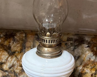 May include: An antique oil lamp with a clear glass chimney and a white, opaque glass reservoir. The lamp has a brass base and accents, showcasing a vintage design. The lamp is a decorative item.