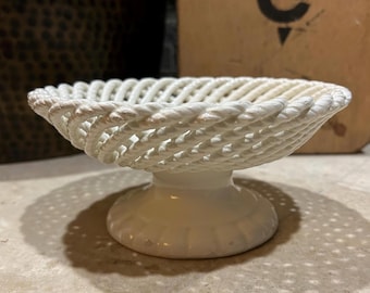 May include: A white ceramic pedestal bowl featuring a woven lattice design along the rim. The bowl has a smooth, solid base and a circular, open-work top that resembles braided rope, creating a textured, interconnected pattern throughout the structure.