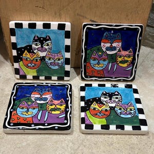 May include: Set of four square ceramic coasters. Each coaster features a colorful, whimsical illustration of three cats with unique patterns and colors, framed by a black and white checkered border. The cat's faces are painted in bright colors.