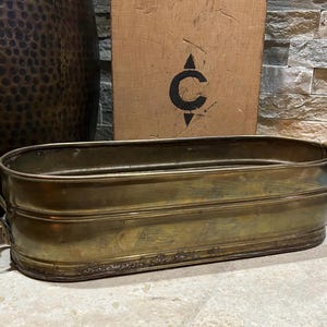 May include: An antique brass planter with an oval shape and ornate handles. The planter has a weathered appearance, with visible signs of age and use. The brass has a warm, golden hue, and the planter is likely used for decorative purposes.