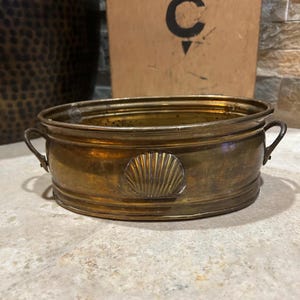 May include: An oval brass basin featuring a polished metallic finish, two small side handles, and a prominent embossed scallop shell motif centered on the front exterior wall. The vessel has a smooth, rounded rim and a deep, open interior cavity.