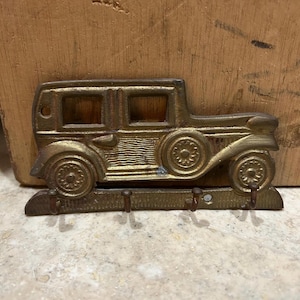 May include: A brass-toned metal wall hook rack shaped like a vintage automobile. The design features a side profile of a car with two windows, a spare tire on the side, and four small hooks extending from the base for hanging items.