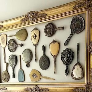 May include: A collection of antique hand mirrors in various shapes and designs, displayed within a gold ornate framed shadow box. The mirrors feature intricate detailing and range in color from silver to ivory.