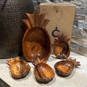 May include: A set of five wooden pineapple-shaped bowls. The bowls are various sizes, with a large bowl in the center and four smaller bowls around it. The wood has a warm, brown tone. The bowls are empty.