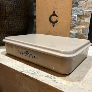 May include: A rectangular beige baking dish with a glossy finish and a blue floral pattern on the side. The dish features a matching lid and shows minor signs of wear along the edges, including small white paint chips on the lower corner.