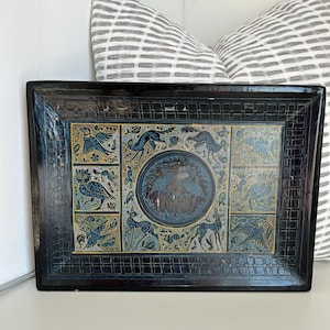 May include: A rectangular, black decorative tray with an intricate, embossed design. The center features a circular motif with birds and animals in blue and gold. The border has a grid-like pattern, adding to the tray's ornate appearance.