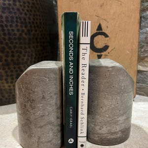 May include: Two heavy, cylindrical concrete bookends with a weathered, grey stone-like texture. Between them stand two books: a dark green spine titled Seconds and Inches by Carly Israel and a white spine titled The Reader by Bernhard Schlink.