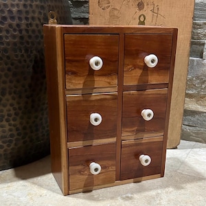 May include: A small, wooden cabinet with six drawers. The cabinet is made of dark brown wood and has white, round knobs. The cabinet is a storage unit, likely for small items or jewelry.