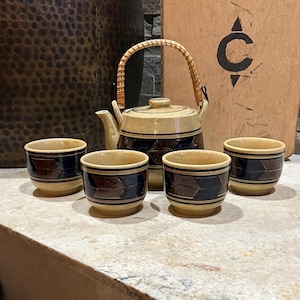 May include: A ceramic tea set with a teapot and four matching cups. The teapot has a woven handle and a beige and brown color scheme. The cups and teapot feature a geometric design with brown and navy blue accents. The set is designed for tea.
