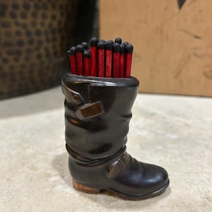 May include: A dark brown boot-shaped match holder filled with red-tipped matches. The boot has buckle details and a worn appearance, suggesting a vintage or rustic style. The matches are neatly arranged, ready for use.