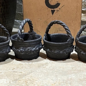 May include: Four small, dark gray cast iron baskets with twisted rope handles. The baskets have a textured surface and decorative floral detailing around the base. They are arranged in a row on a stone surface.
