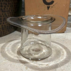 May include: A clear glass vessel shaped like a top hat with a wide, textured brim. The cylindrical body is smooth and transparent, while the outer edge of the brim features a distinct, frosted, or stippled surface texture that catches the light.