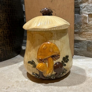 May include: A ceramic mushroom-shaped jar with a lid. The jar is beige with a brown lid and a cluster of orange mushrooms on the front. Green leaves are also present. The jar is likely used for storage.