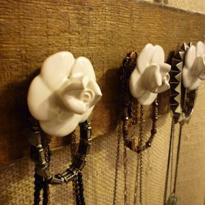 May include: White ceramic rose-shaped hooks mounted on a wooden board with multiple necklaces hanging from them.