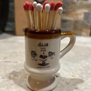 May include: A small ceramic mug filled with wooden matches. The matches have red or white tips. The mug is beige with a brown rim and handle, and features illustrations and text related to Ohio, including "Cleveland" and "Columbus State Capitol."