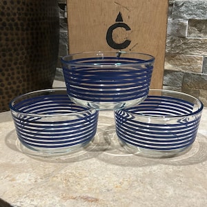 May include: Three clear glass bowls with horizontal blue and white stripes. The bowls are stacked on a light-colored surface. A wooden board with a black symbol is in the background.