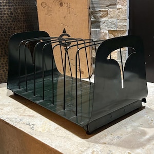 May include: Dark green metal magazine rack with a series of vertical dividers. The rack has a rectangular base and curved sides with a decorative cutout. The dividers are made of thin, black metal rods.