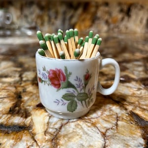 May include: A small, white teacup filled with green-tipped matches. The cup is decorated with a floral pattern featuring red roses and green leaves. The cup has a small handle and is set on a patterned surface.