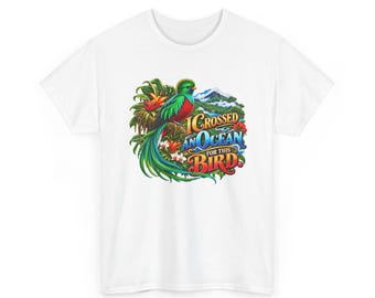 Pode incluir: Uma t-shirt branca com um gráfico colorido de um quetzal pousado em folhagem tropical com uma paisagem montanhosa ao fundo. O texto diz I CROSSED AN OCEAN FOR THIS BIRD em letras estilizadas azuis e vermelhas.