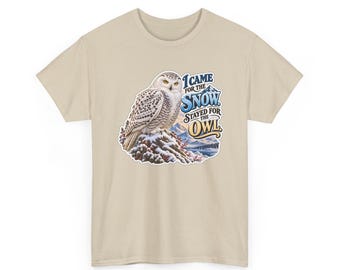 Pode incluir: Uma t-shirt bege com um gráfico de uma coruja-das-neves pousada numa rocha coberta de neve. O texto diz I CAME FOR THE SNOW STAYED FOR THE OWL, acompanhado por uma ilustração de uma paisagem montanhosa em tons de azul e branco.