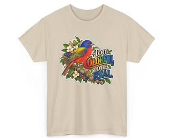 Pode incluir: Uma t-shirt bege de manga curta com o gráfico de um pássaro multicolorido pousado num ramo com flores brancas e folhas verdes. O texto "TOO COLORFUL TO BE REAL" está impresso em letras com as cores do arco-íris ao lado do pássaro.