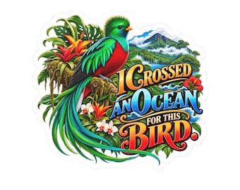 Pode incluir: Um adesivo recortado com um quetzal de cores verde e vermelho vibrantes pousado num ramo entre folhagem e flores tropicais. Ao fundo, uma montanha ergue-se sobre um oceano azul. O texto diz I CROSSED AN OCEAN FOR THIS BIRD.