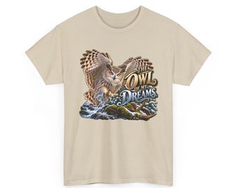 Pode incluir: Uma t-shirt bege de manga curta com um gráfico de uma coruja castanha com as asas abertas a voar sobre um riacho rochoso. O texto "THE OWL OF MY DREAMS" está impresso numa fonte estilizada ao lado da coruja, com fundo de floresta.