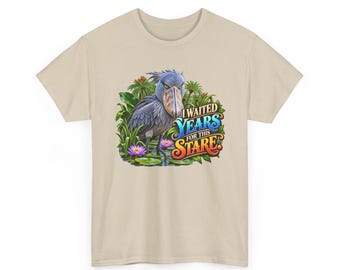 Pode incluir: Uma t-shirt bege com um gráfico de um bico-de-tamanco em pé num lago com plantas verdes e nenúfares roxos. O texto "I WAITED YEARS FOR THIS STARE" está impresso numa fonte colorida e estilizada ao lado da ilustração da ave.