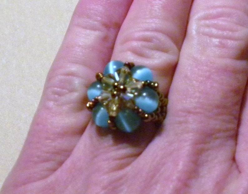May include: A ring featuring a flower design crafted from blue and gold beads. The ring is worn on a finger.