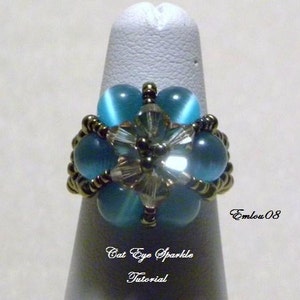 May include: A ring with a blue cat's eye stone center surrounded by smaller blue stones and silver beads. The ring is on a white display stand. The text "Cat Eye Sparkle Tutorial" and "EmLou 08" are visible on the stand.