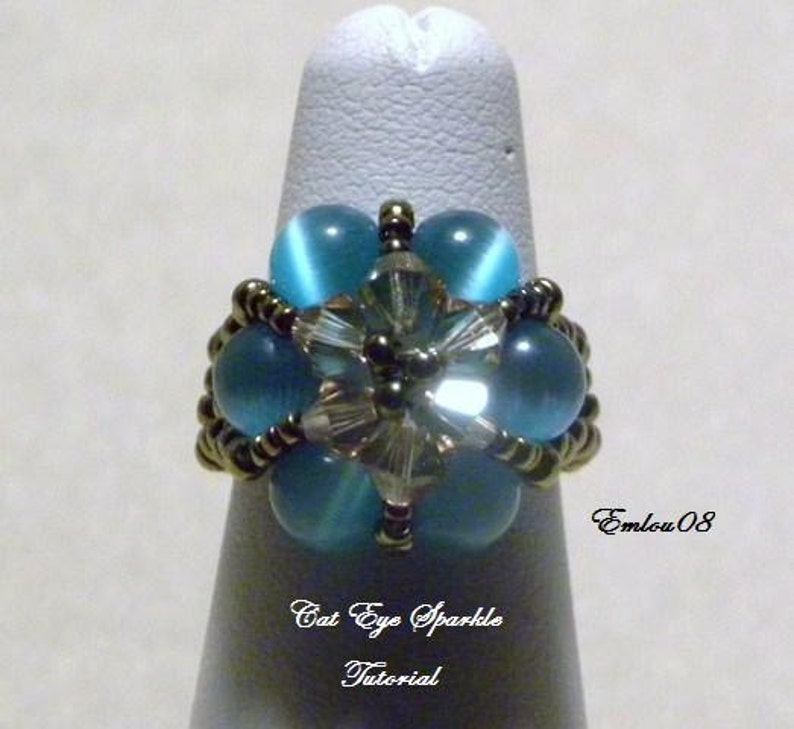 May include: A ring featuring a blue cat's eye stone centre surrounded by smaller blue stones and silver beads. The ring is displayed on a white stand. The text "Cat Eye Sparkle Tutorial" and "EmLou 08" are visible on the stand.