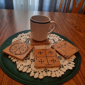 May include: Four wooden coasters with different designs. The coasters are on a white doily on a green round placemat. A white mug with a blue band is sitting on one of the coasters.