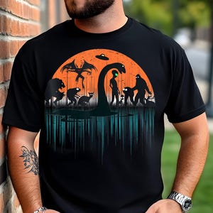 Könnte beinhalten: Ein schwarzes T-Shirt mit einem grafischen Druck eines Sonnenuntergangs mit verschiedenen mythischen Kreaturen, darunter ein Monster, ein Bigfoot und ein Außerirdischer. Die Kreaturen sind vor einem orangefarbenen Sonnenuntergang mit einer fliegenden Untertasse darüber silhouettiert.