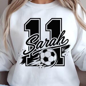 May include: A white crewneck sweatshirt featuring a large black graphic of the number 11 with a white outline. The name Sarah is written in a black script font across the center, layered over a black and white soccer ball with brush stroke accents.